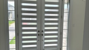 Eine Doppeltür mit horizontalen Lamellen, eine schwarze Fußmatte und Blick auf eine Wohnstraße.