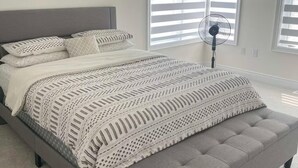 Ein Schlafzimmer mit einem grau gepolsterten Bett, weißer Bettwäsche und einem Standventilator.
