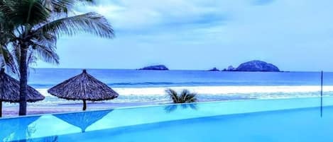 Enjoy a tranquil beachfront setting with a clear ocean view, a swimming pool, and shaded areas under thatched umbrellas.