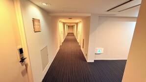 Een hotelgang met vloerbedekking, een deur aan de linkerkant en een bord met telefoonnummers aan de muur.