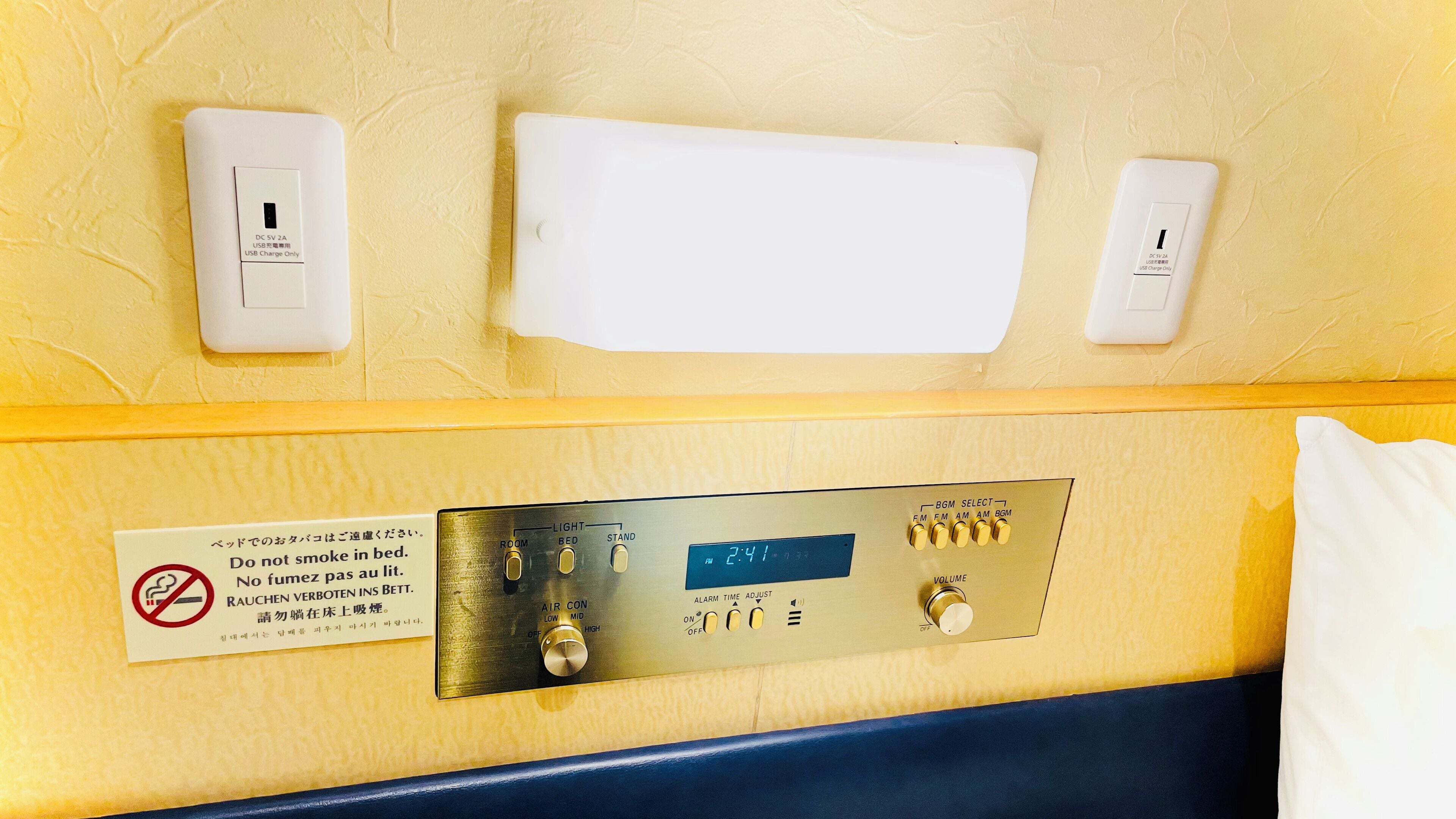 A hotel room with a bedside table, a digital clock, and a sign in a foreign language.