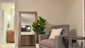 Ein Wohnzimmer mit einem grauen Sessel, einem runden Holztisch, einem großen Spiegel, einem Fernseher und einer Holzkommode.