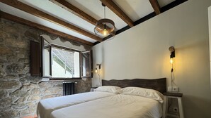 A bedroom with a stone wall, wooden beams, a large bed, a nightstand, and a window with a view. - La Melera - Starlight lodging and skiing (Llanos)