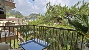 Un balcon doté d’une table à plateau de verre et d’un canapé, entouré d’une rambarde en métal, offrant une vue sur un paysage verdoyant avec des arbres et des bâtiments.
