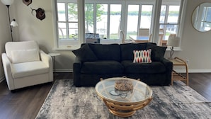 A living room with a dark green sofa, a white armchair, and a glass coffee table with a woven base. - Fully renovated lake front home
on Delavan’s North Shore (Delavan)