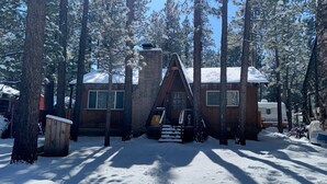A cabin with a brick chimney, surrounded by tall pine trees and covered in snow.