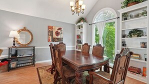 Comedor con mesa y sillas de madera, candelabro, estantería y un ventanal con vistas a la vegetación.