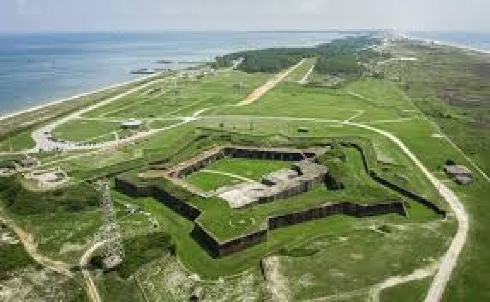 A large, open fort-like structure with multiple walls and pathways, surrounded by green fields and a coastal view.
