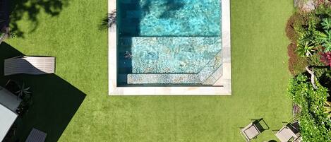 Vista aérea de una piscina rectangular rodeada de césped verde, sillas de madera y plantas tropicales.