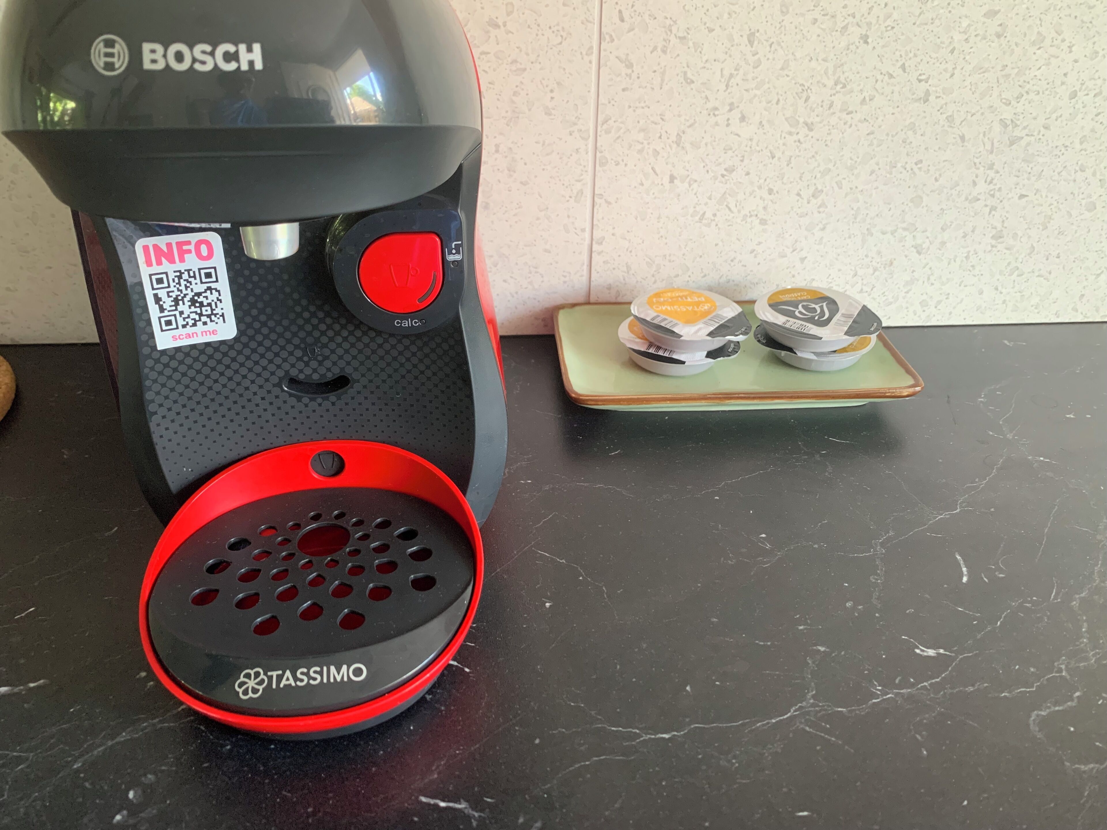 Een Bosch koffiezetapparaat met een rode basis en een Tassimo houder voor capsules, geplaatst op een donker aanrecht naast een groene dienblad met twee koffiecapsules.