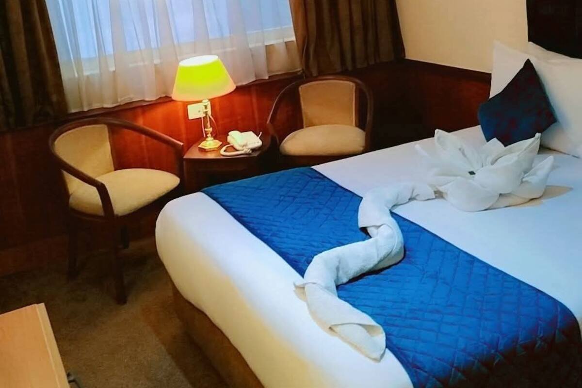 A hotel room with a large bed, a desk with a lamp, a chair, and a window with curtains.