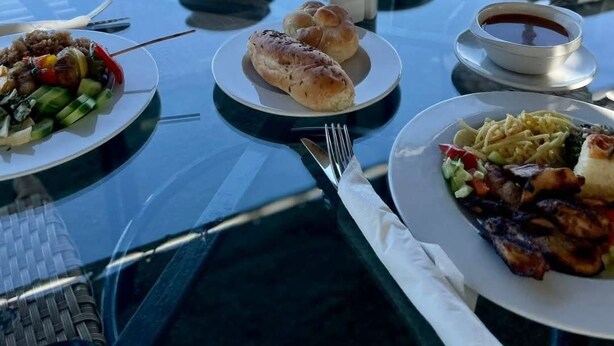 Une table dressée pour un repas, avec différents plats, notamment de la viande grillée, des légumes et du pain, donnant sur une étendue d’eau.