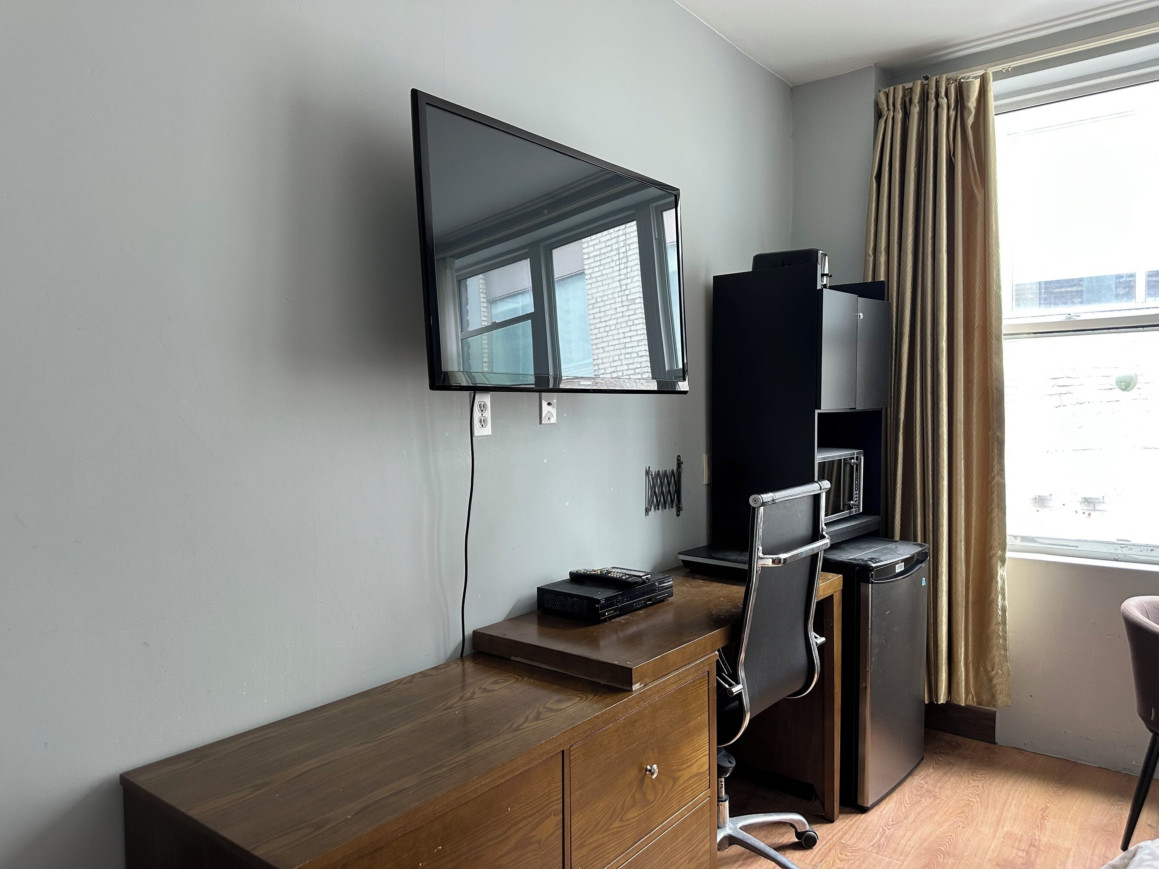 A hotel room with a wooden desk, a flat-screen TV mounted on the wall, a refrigerator, and a window with curtains.