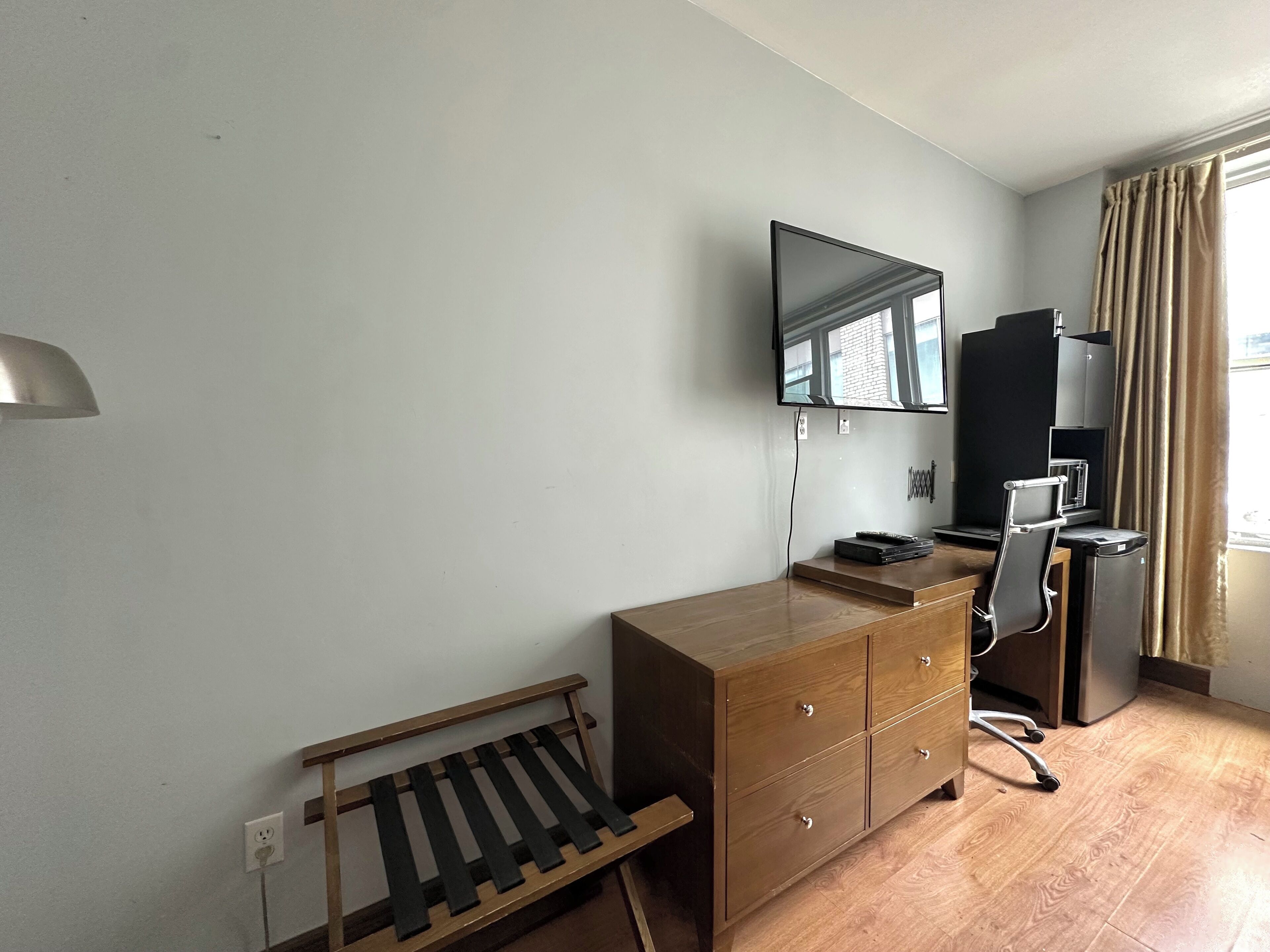 Quarto compacto com penteadeira de madeira, escrivaninha, cadeira, televisão fixada na parede e janela com cortinas.
