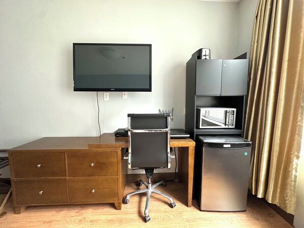 Una camera d'albergo con una scrivania in legno, una sedia da ufficio nera, una TV a schermo piatto fissata al muro, un frigorifero e un microonde.