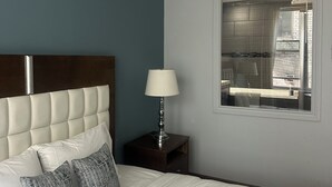A hotel room with a bed, a nightstand, a lamp, and a window with a view of the city. - King bed suite in the heart of downtown, festive atmosphere (Montréal)
