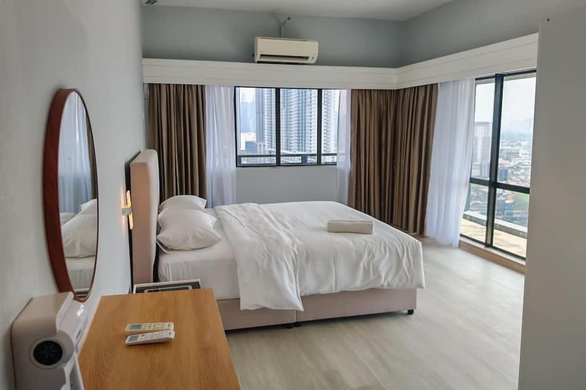 Quarto de hotel com cama, escrivaninha, televisor remoto, espelho e janela com cortinas.