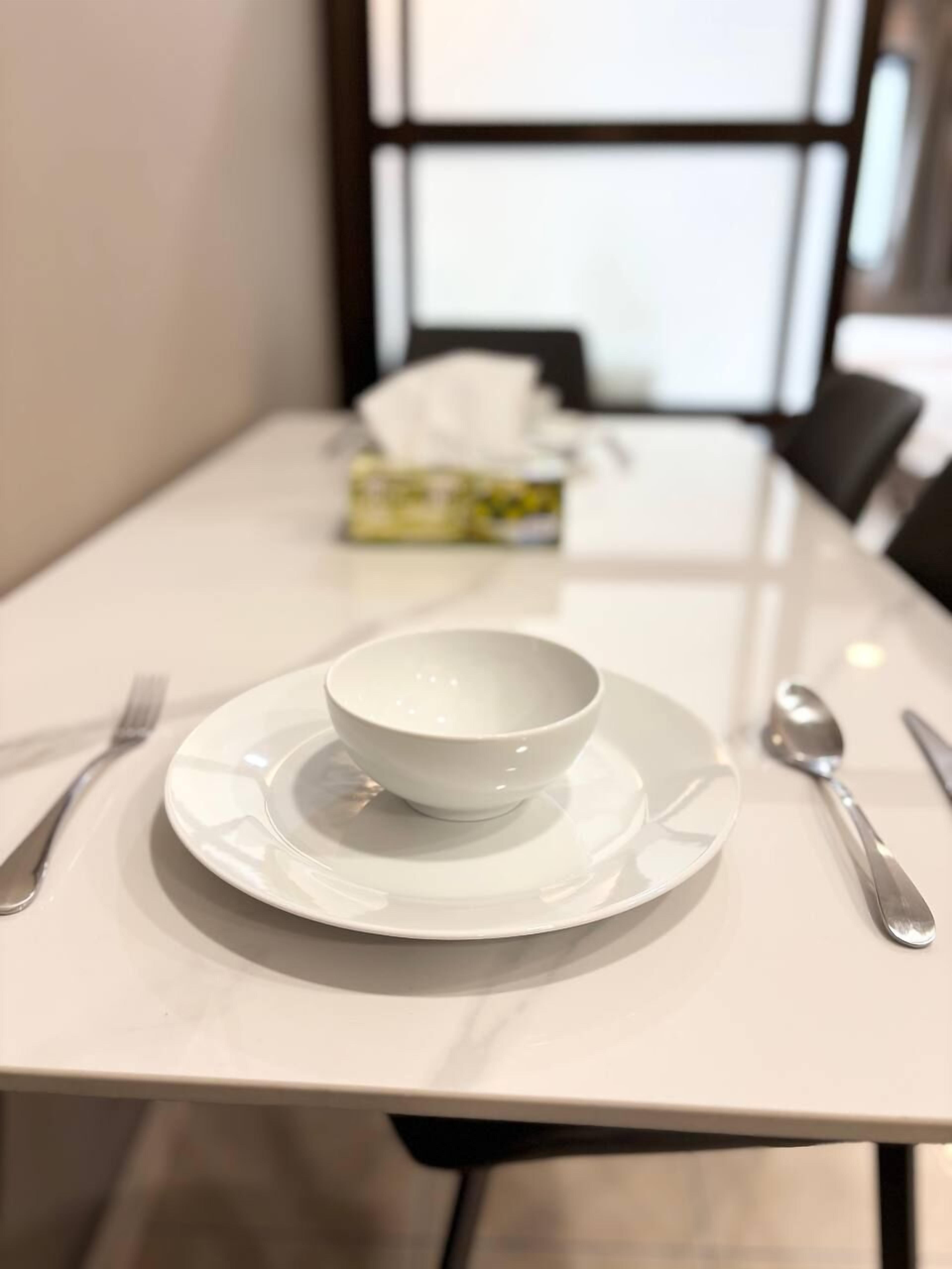 A dining table with a white bowl and plate, silverware, and a napkin dispenser in the background.