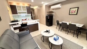 A modern living room with a grey sofa, two round coffee tables, a dining area with a table and chairs, a kitchen with cabinets and appliances, and an air conditioning unit on the wall. - Cempaka Apartment By Easy Nap (Kuala Lumpur)