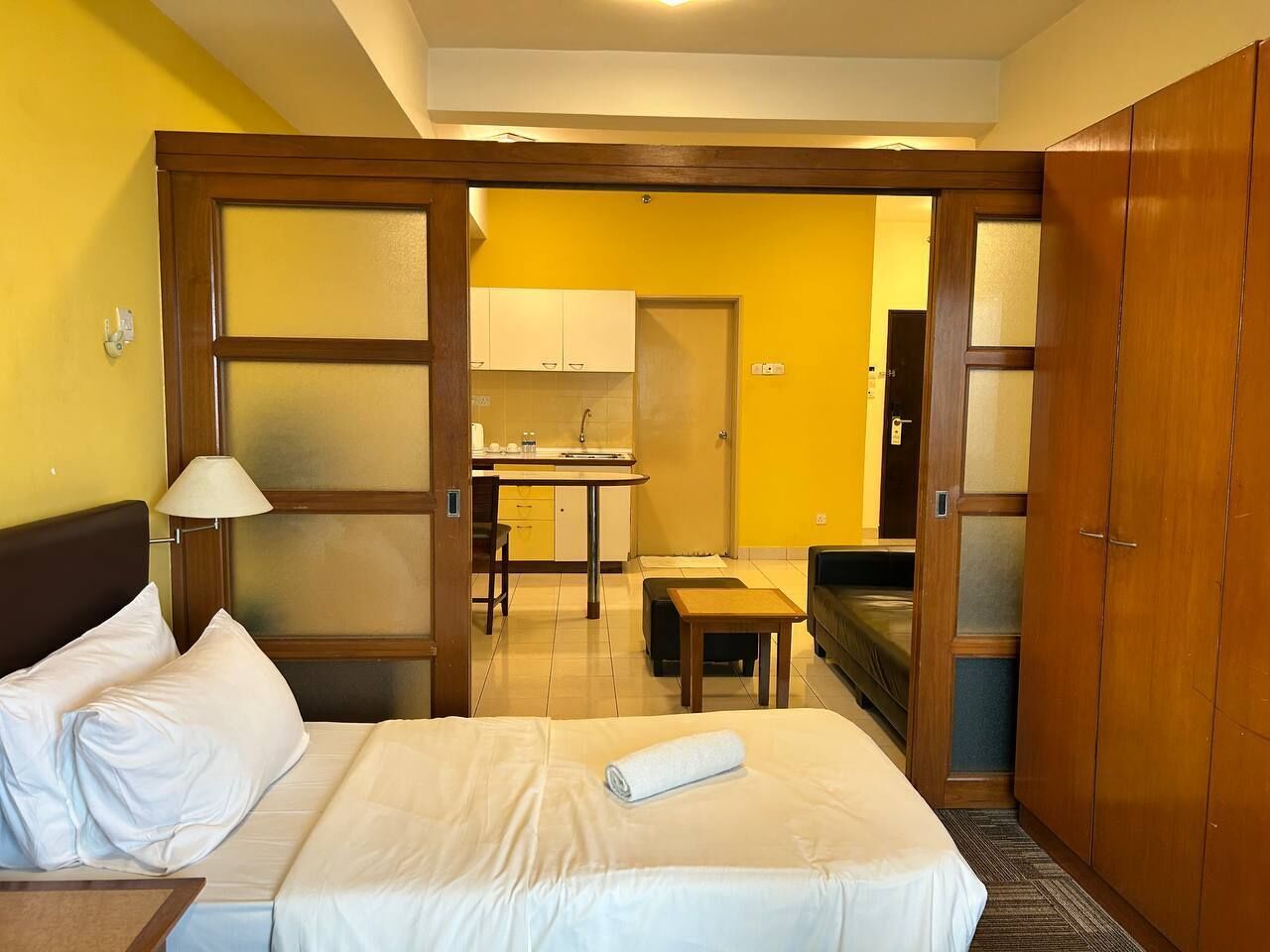 A hotel room with a bed, bedside table, and a wooden wardrobe. There is a kitchenette in the background.