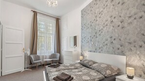 A bedroom with a large bed, a bedside table with a lamp, a chair, a window with curtains, and a chandelier.