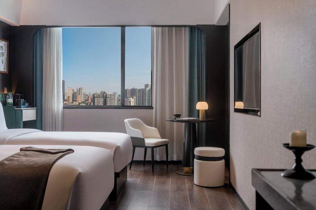 A hotel room with two beds, a small table with a lamp, a chair, and a view of the city skyline.