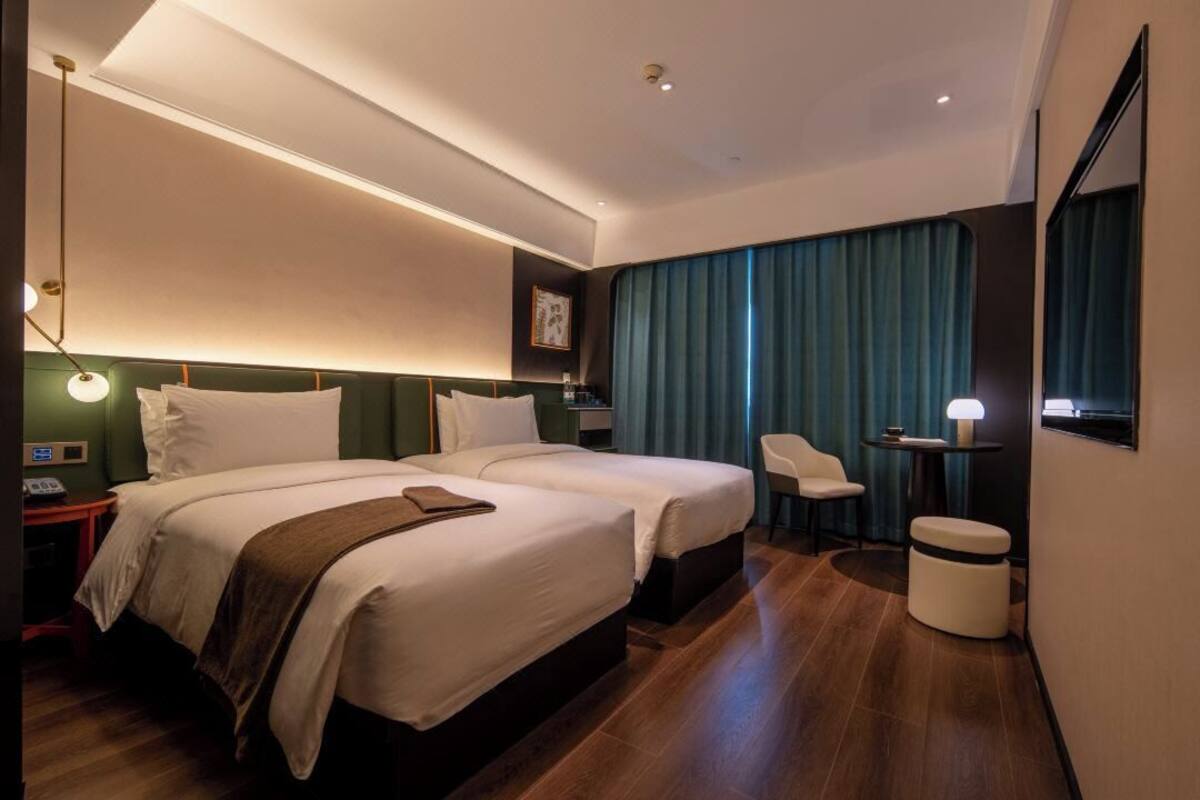 Ein modernes Hotelzimmer mit zwei Betten, einem Schreibtisch, einem Stuhl und einem kleinen Tisch mit einer Lampe.