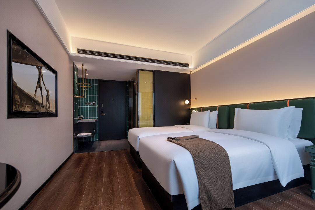 Ein modernes Hotelzimmer mit einem großen Bett, einem Nachttisch, einem gerahmten Bild an der Wand, einem Badezimmer mit Glasabtrennung und Holzboden.