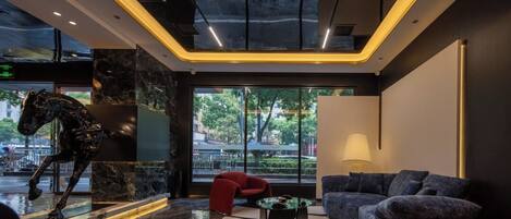 Un vestíbulo moderno con piso de mármol, una escultura, una silla roja, un sofá azul y un amplio ventanal con vistas al exterior.