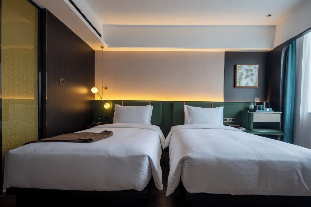 A hotel room with two single beds, a nightstand with a lamp, a small table with a cup, and a framed picture on the wall.