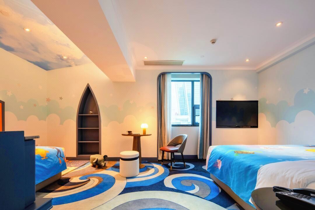Ein Hotelzimmer mit zwei Betten, einem Fernseher, einem Schreibtisch, einem Stuhl und einem Fenster mit Blick auf Gebäude.