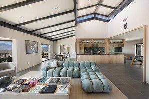 A modern living room with a sectional sofa, a dining area, and a kitchen with wooden cabinets. - Addendum Estate - 2 Pools, Panoramic Views & Luxe Privacy for Groups (Desert Hot Springs)
