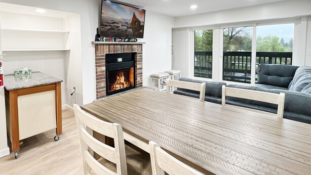 A modern living room with a large wooden dining table, a fireplace, a television, and a view of the outdoors. - Newly Remodeled 1BR | Shuttle, Fireplace, Amenities in the Heart of Killington (Killington)
