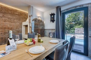 A dining area with a wooden table set for four, a brick wall, a stone fireplace, and a view of a pool and greenery. - Stone Villa Galant with Pool, Brgudac, Croatia (Brgudac)