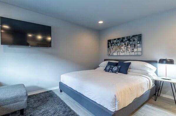 A modern bedroom with a large bed, a flat-screen TV mounted on the wall, a small bedside table with a lamp, and a gray armchair. - Montrose Chic Studio! Medical Center 12 (Houston)