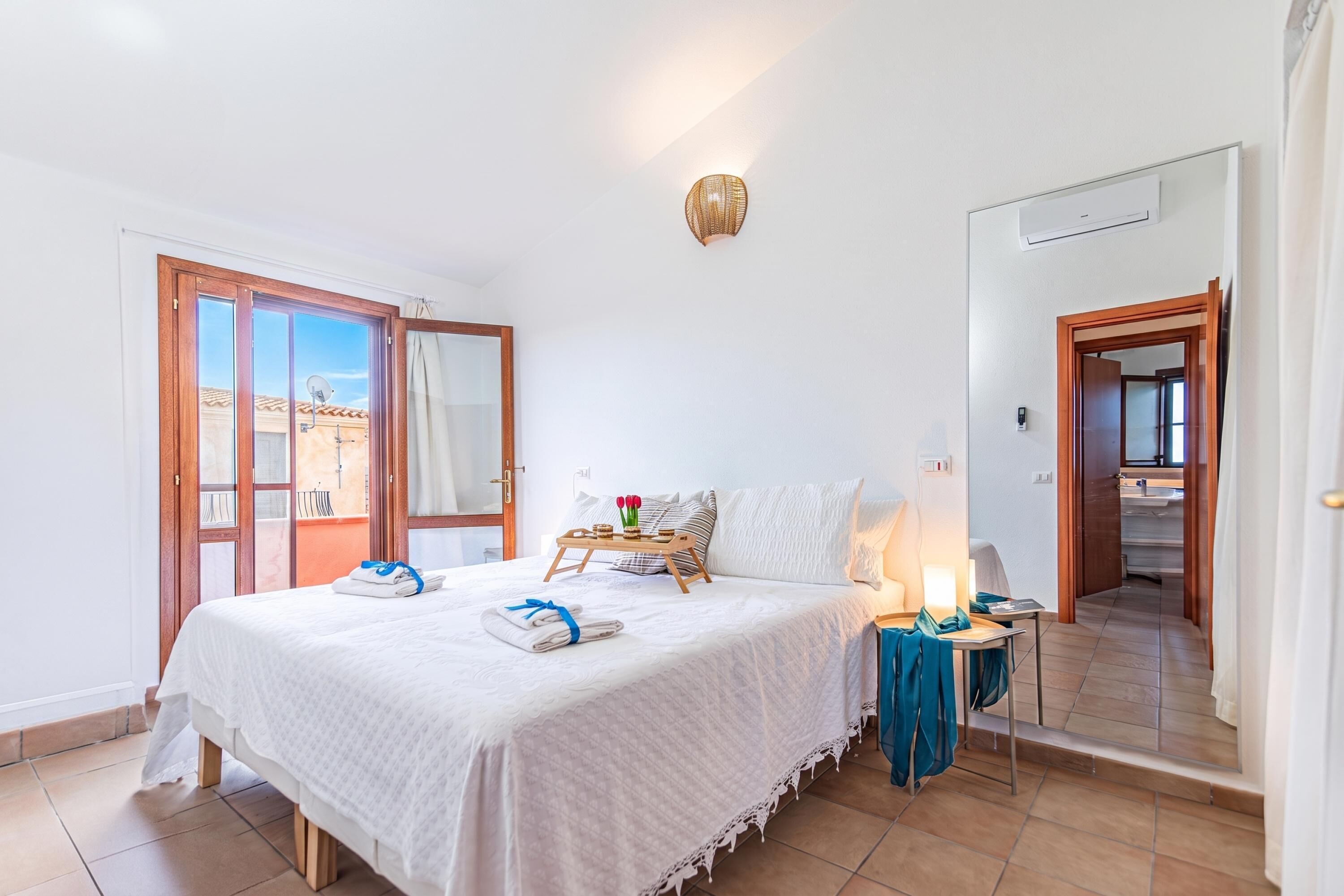 A hotel room with a bed, a bedside table, a lamp, a window with a view, and a door leading to the bathroom.