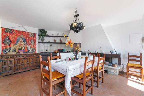 A dining area with a table set for six, surrounded by wooden chairs with yellow cushions. A tapestry hangs on the wall, and a hanging light fixture is visible above the table.