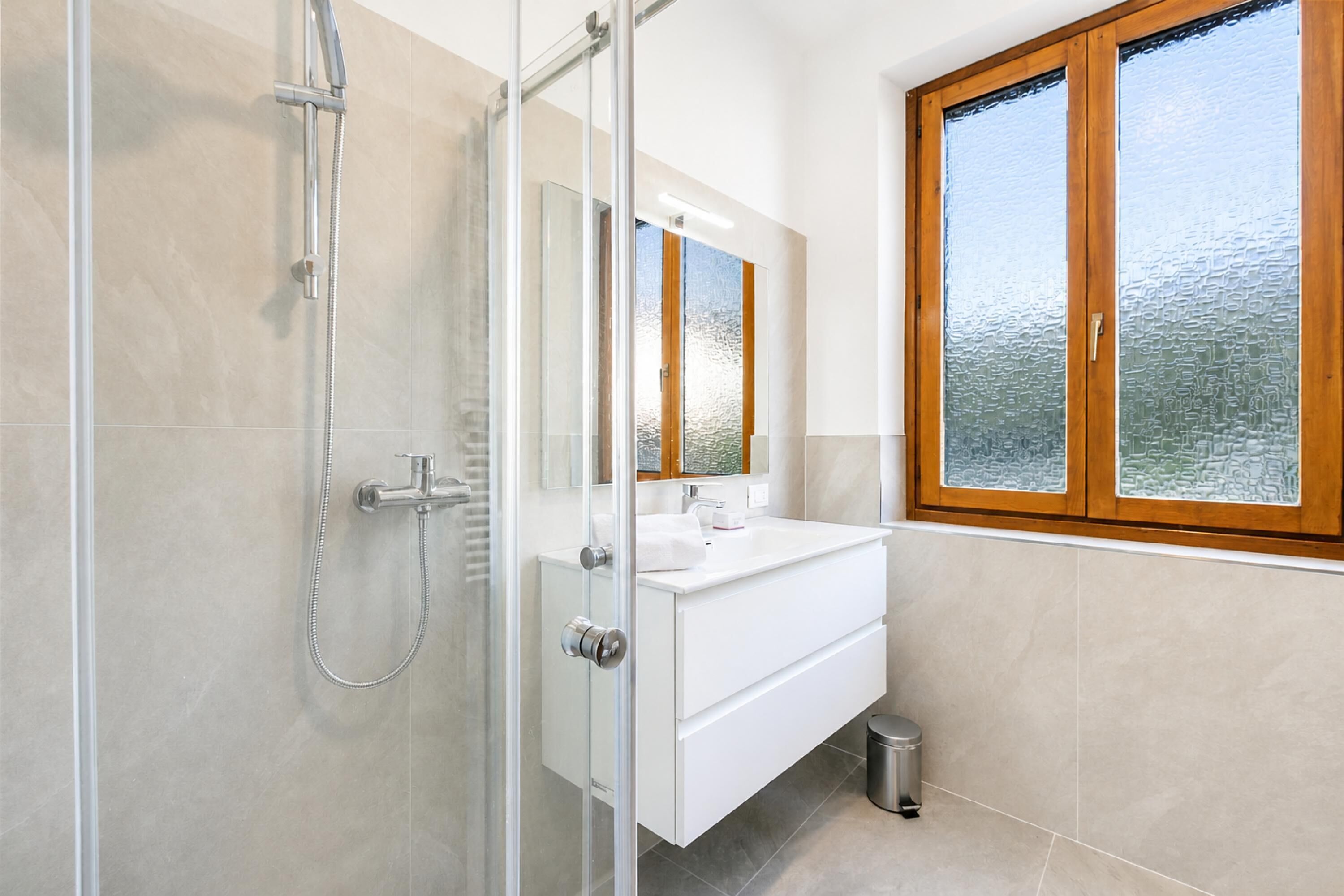 A modern bathroom with a glass shower enclosure, a white sink, and a large window with wooden frames.