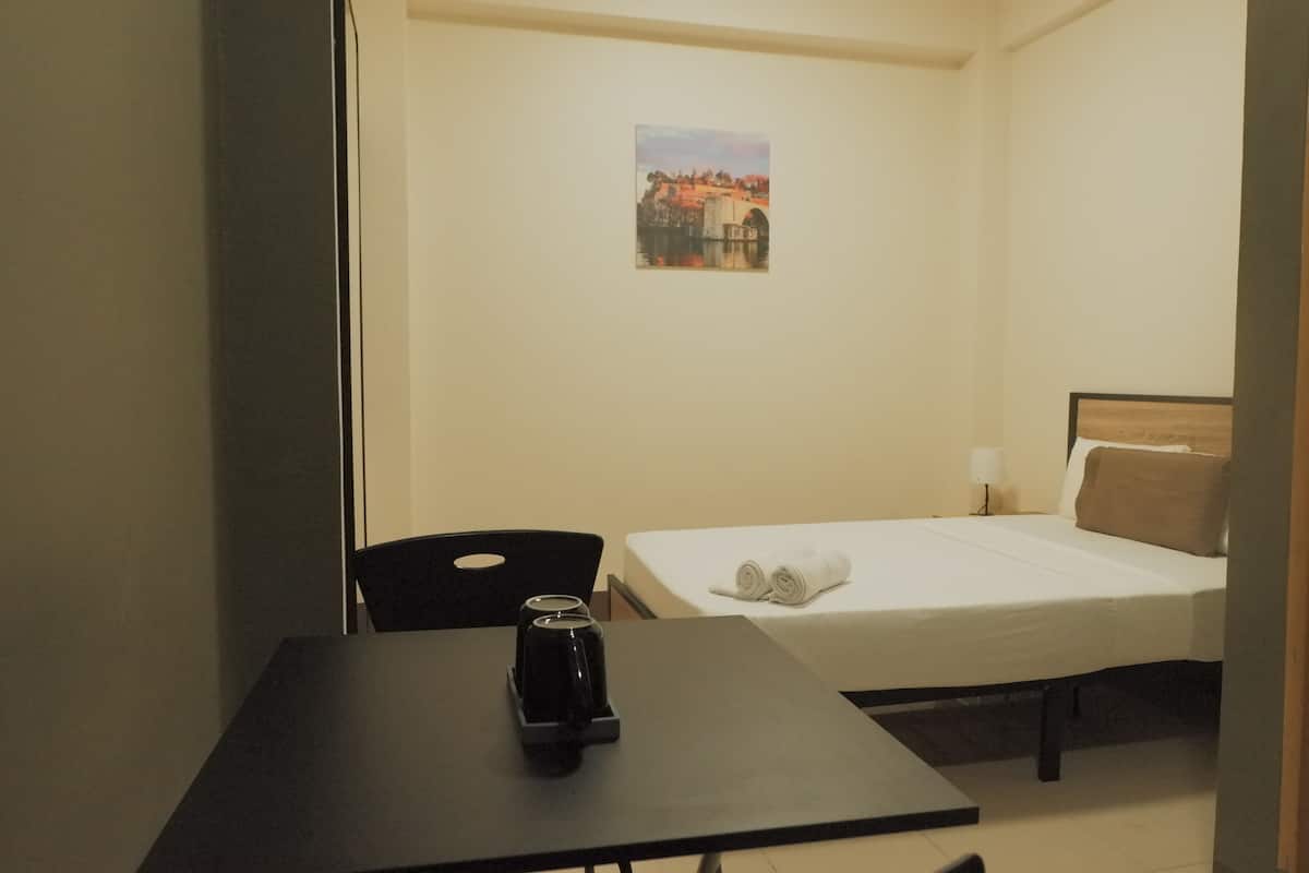 Een kleine, verzorgde kamer met een eenpersoonsbed, een tafel met waterkoker en een aan de muur hangend schilderij.