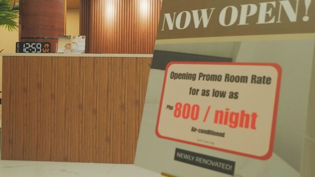 A hotel reception desk with a digital clock displaying 12:59 and a promotional sign for an opening promo room rate of 800 per night. - Brightwood Hotel & Residences (Manila)