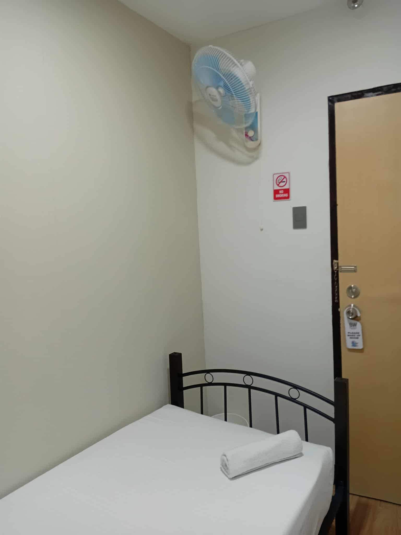 Una stanza piccola con un letto singolo, una ventola a muro, una porta con serratura a chiave card e un cartello che indica divieto di fumo.
