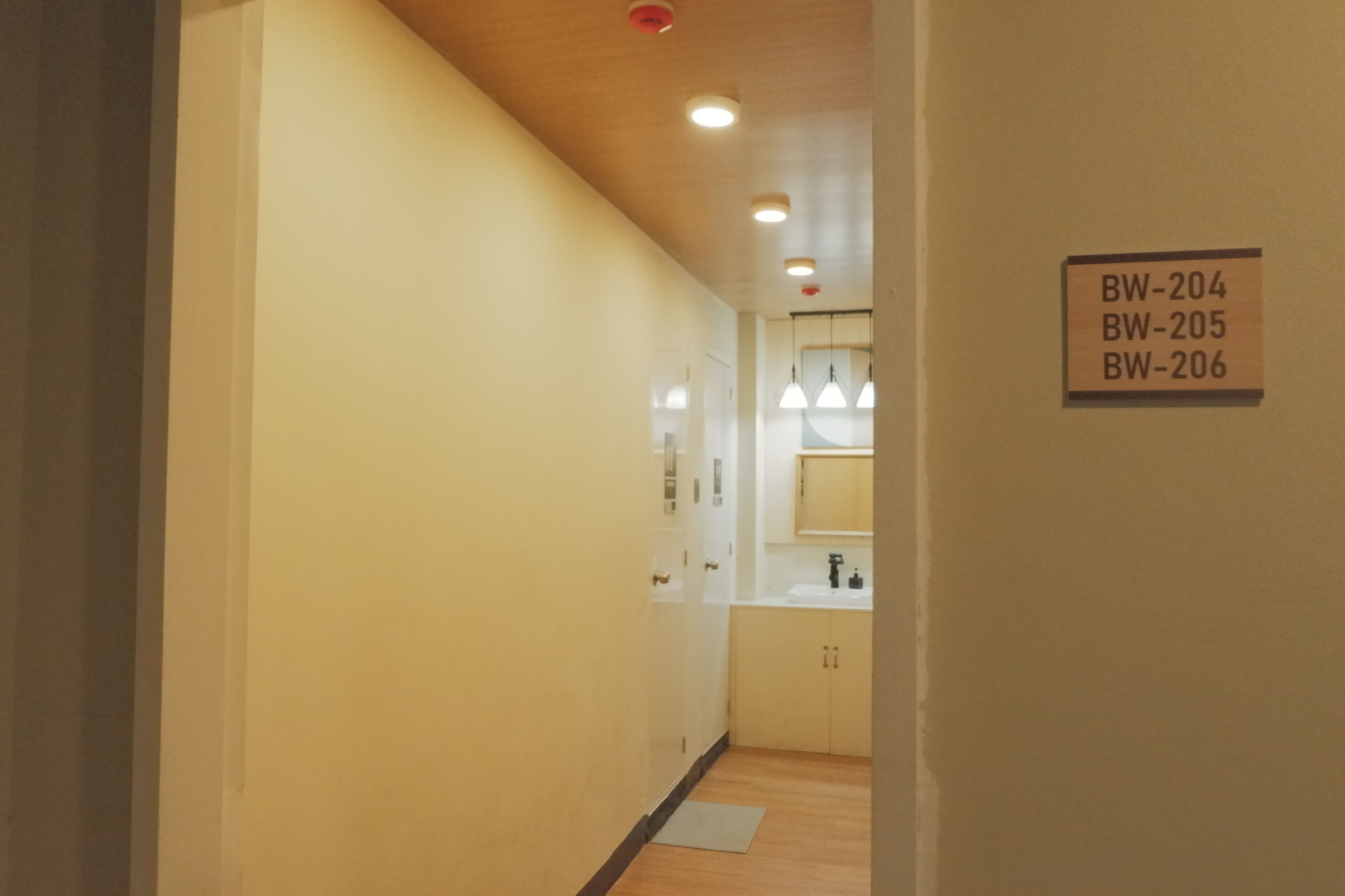 Ein Flur mit Holzboden und weißen Wänden, der zu einem Badezimmer mit Waschbecken und Spiegel führt. Deckenleuchten und ein Schild mit Zimmernummern sind vorhanden.