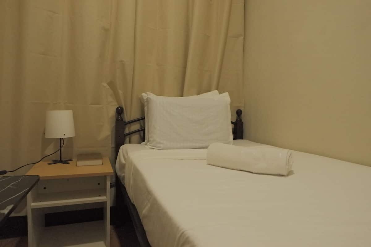 Een net opgemaakt bed met witte lakens, een bedlampje en een klein nachtkastje met een boek en een lamp.