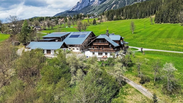 Ein traditionelles Chalet mit geneigtem Dach, umgeben von üppigen grünen Feldern und Bergen im Hintergrund.