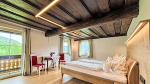 Ein Schlafzimmer mit Holzboden, einem Bett mit floraler Bettwäsche, einem kleinen Tisch mit Stuhl und Blick ins Grüne.