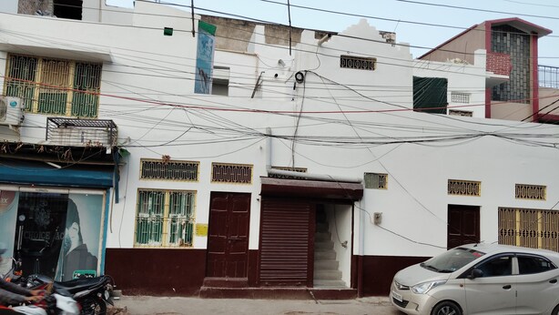 A building with multiple windows, some with metal bars, a closed brown door, and a parked white car. - Ksn guest House  (Varanasi)