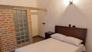 Una cama bien hecha con sábana y almohada blancos, cabecera de madera, una mesita de noche pequeña y una pared de ladrillo con una ventana.