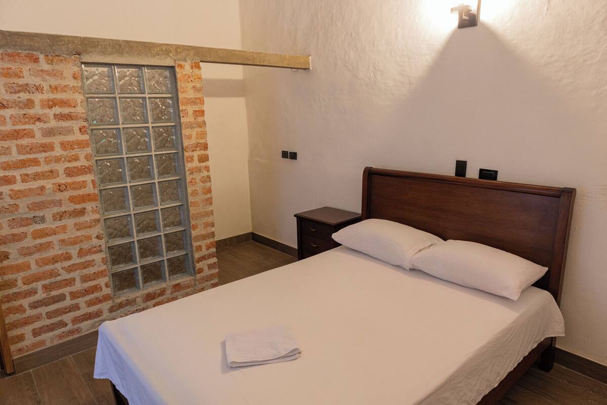 A neatly made bed with a white sheet and pillow, a wooden headboard, a small bedside table, and a brick wall with a window.