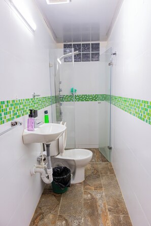Un baño con lavamanos, inodoro y ducha blancos. Las paredes están cubiertas con azulejos blancos y verdes, y hay una ventana sobre la ducha.