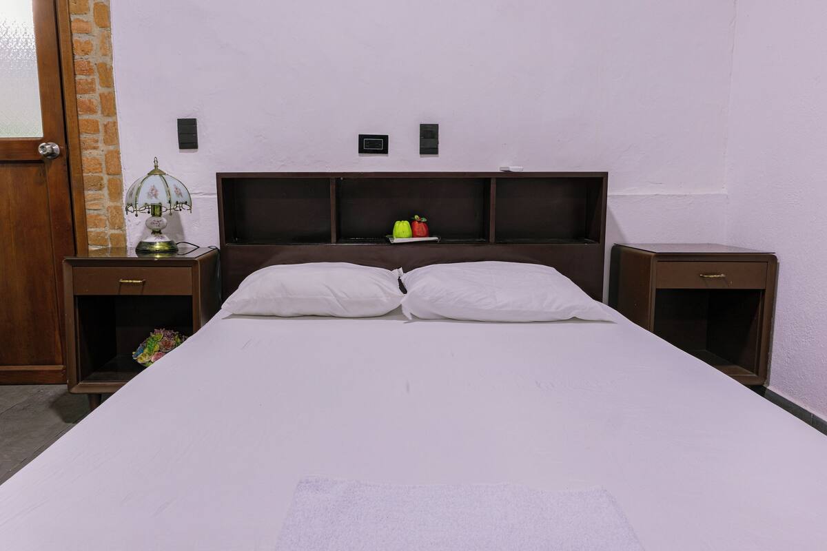 A hotel room with a large bed, two bedside tables, a nightstand, a wall-mounted shelf with decorative items, and a door with a glass panel.